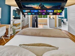 A modern hotel room with a large flat-screen TV displaying a movie, a bed with a beige blanket, a desk with a chair, and a small kitchenette. - Private Studio/1 Min-Hospital/Self Check-In/75" TV (Williams Lake)