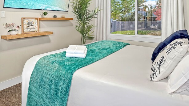 A bedroom with a bed, a TV on the wall, a window with curtains, a plant, and a shelf with decorative items. - Cozy Suite/1 Min-Hospital/Full Kitchen/Sleeps 4 (Williams Lake)