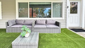 A modern outdoor seating area with a wicker sofa and a matching coffee table, set against a backdrop of a house with large windows and a well-maintained lawn. - Cozy Suite/1 Min-Hospital/Full Kitchen/Sleeps 4 (Williams Lake)