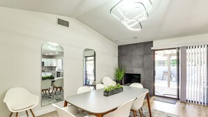 Un espace repas moderne avec une grande table, des chaises blanches, une cheminée et une vue à travers une porte vitrée.