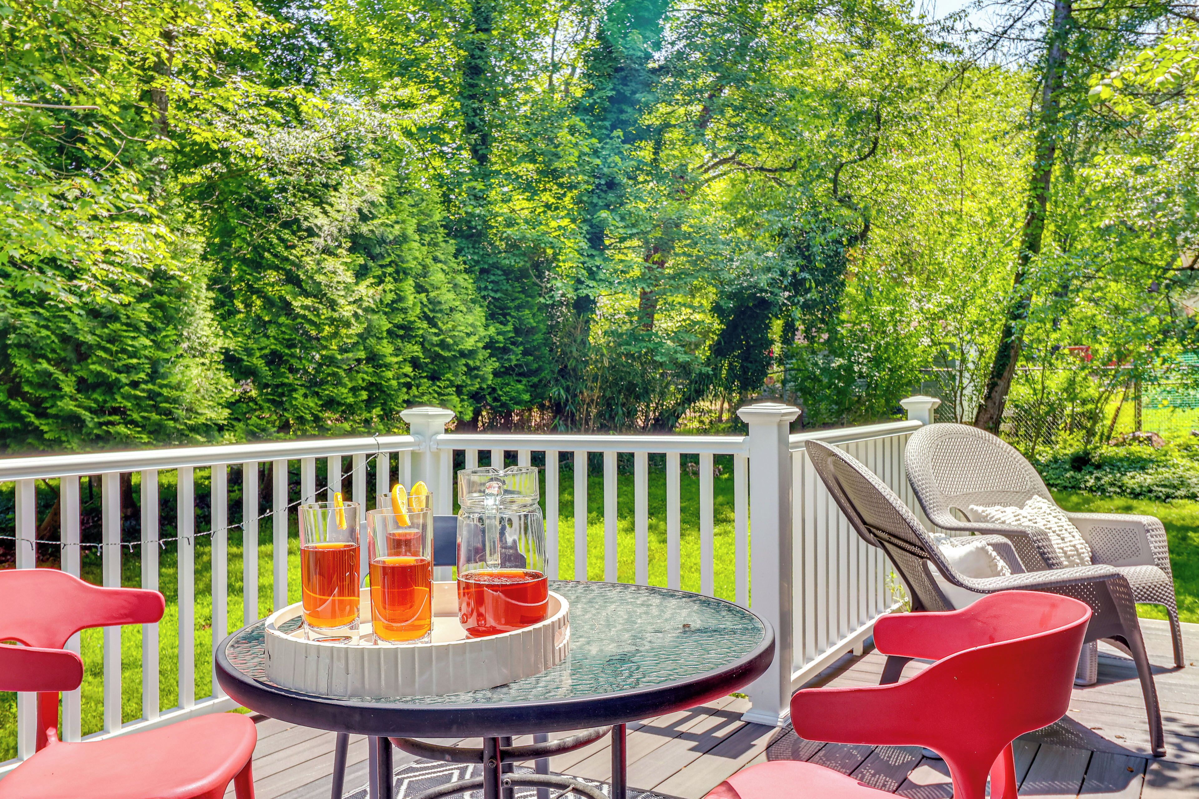 A deck with a round table set for drinks, surrounded by red chairs, overlooking a lush green garden.