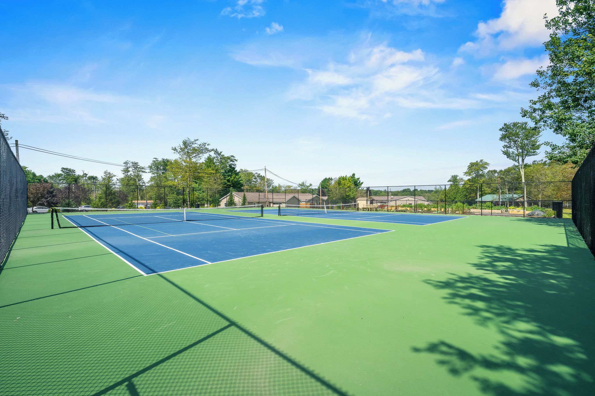 An outdoor tennis court with a green and blue surface, surrounded by trees and buildings.