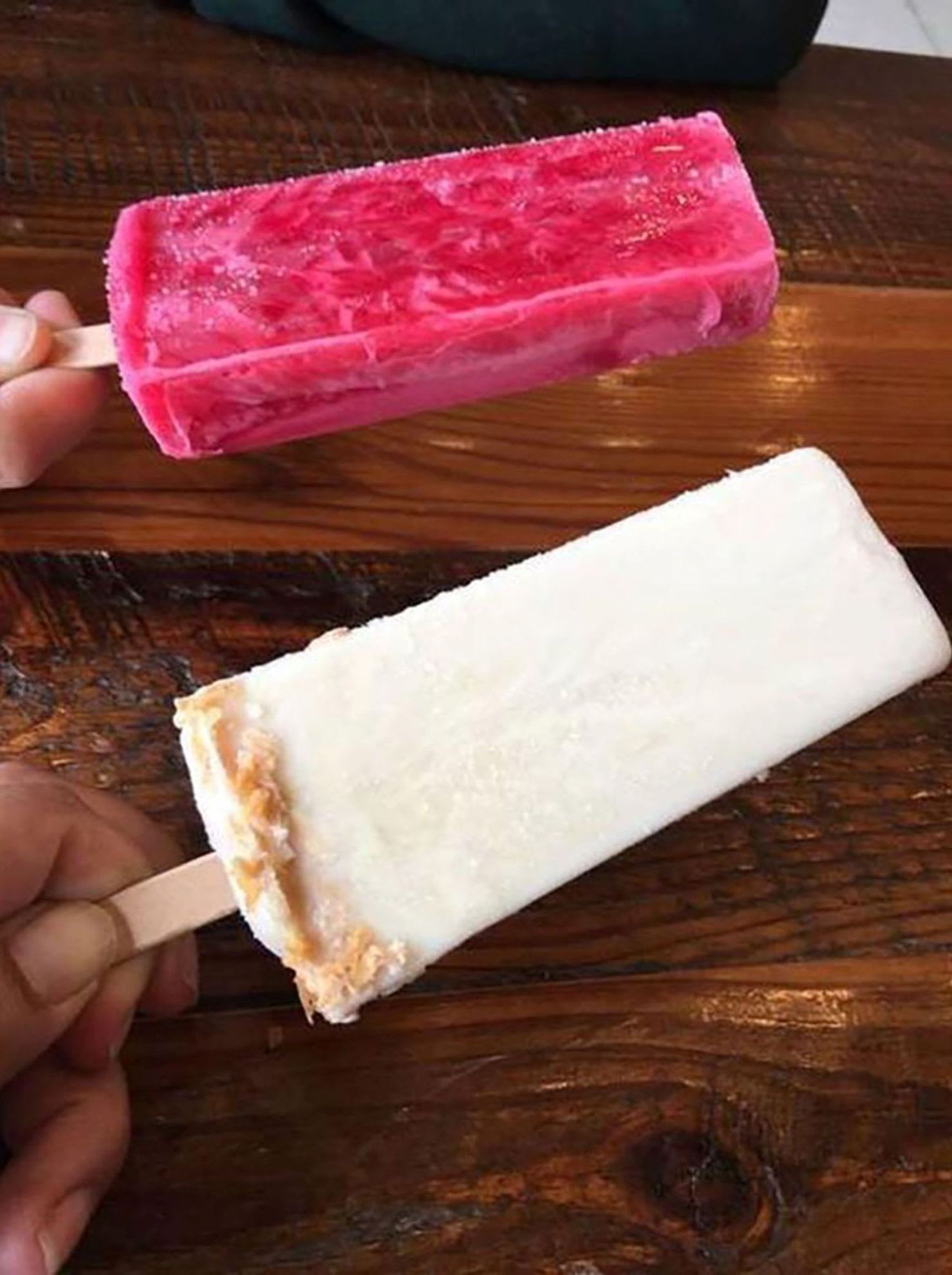 Une table en bois sur laquelle reposent deux bâtonnets de glace, l’un rose et l’autre blanc, tenus dans une main.