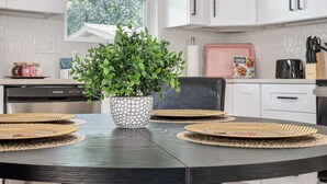 A modern kitchen with white cabinets, a round dining table with woven placemats, a potted plant, and a cooking book on the counter. - Historic Comfort 2BR 1BA Guesthouse Near Riverwalk (San Antonio)