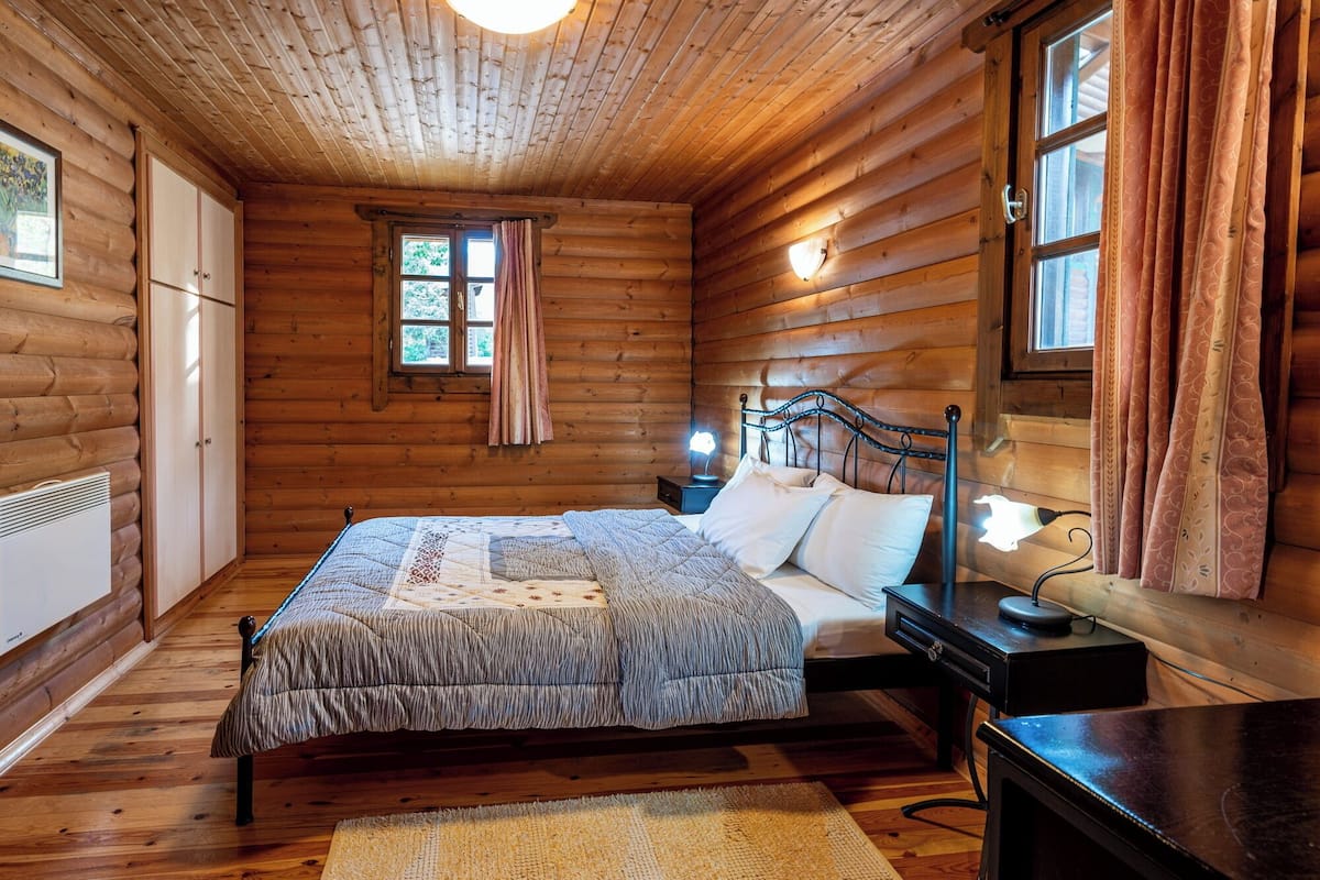 A log cabin bedroom with a bed, bedside tables, a nightstand, a window with curtains, and a radiator.