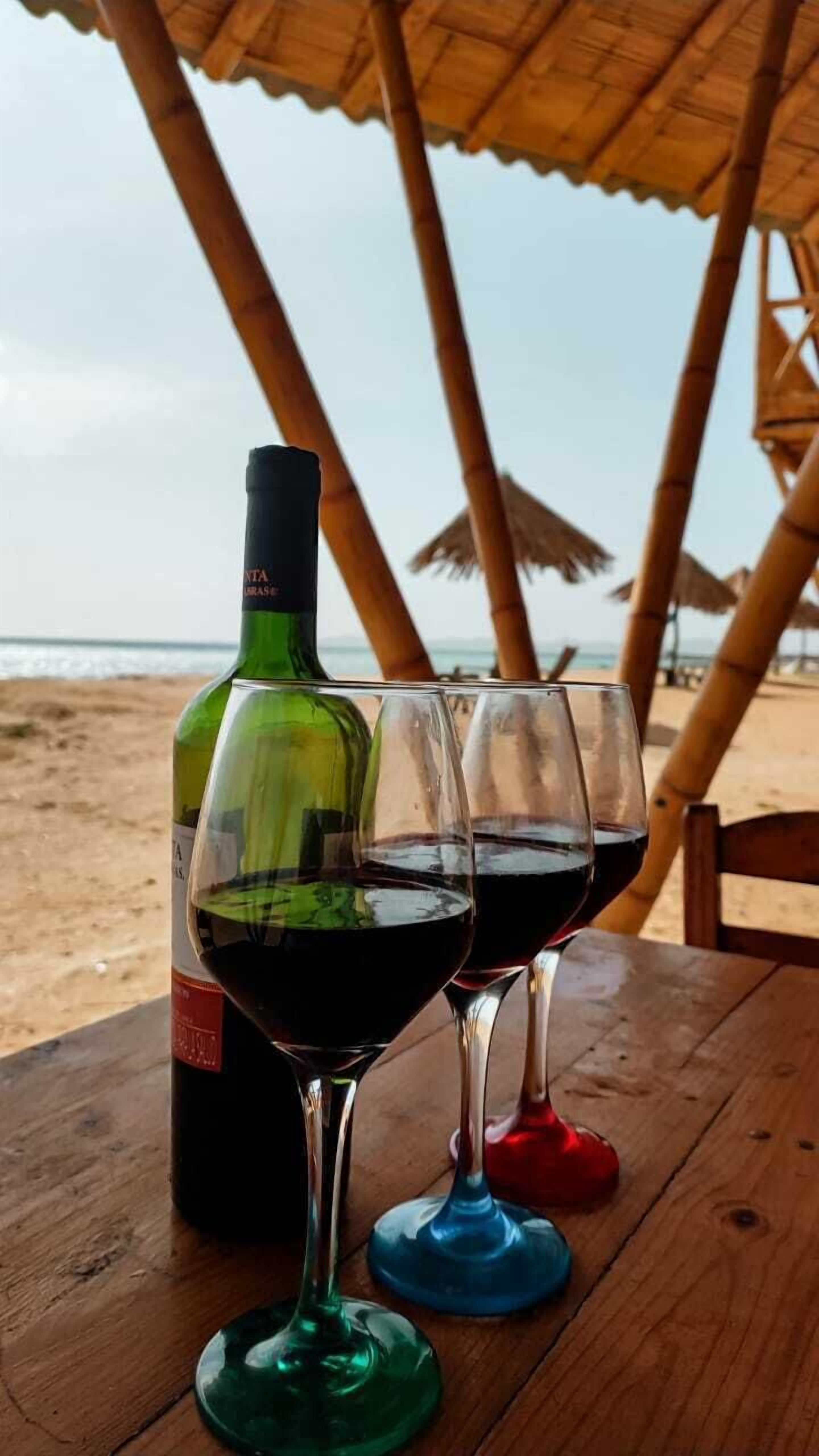 A bottle of wine and three glasses on a wooden table at a beachside location.