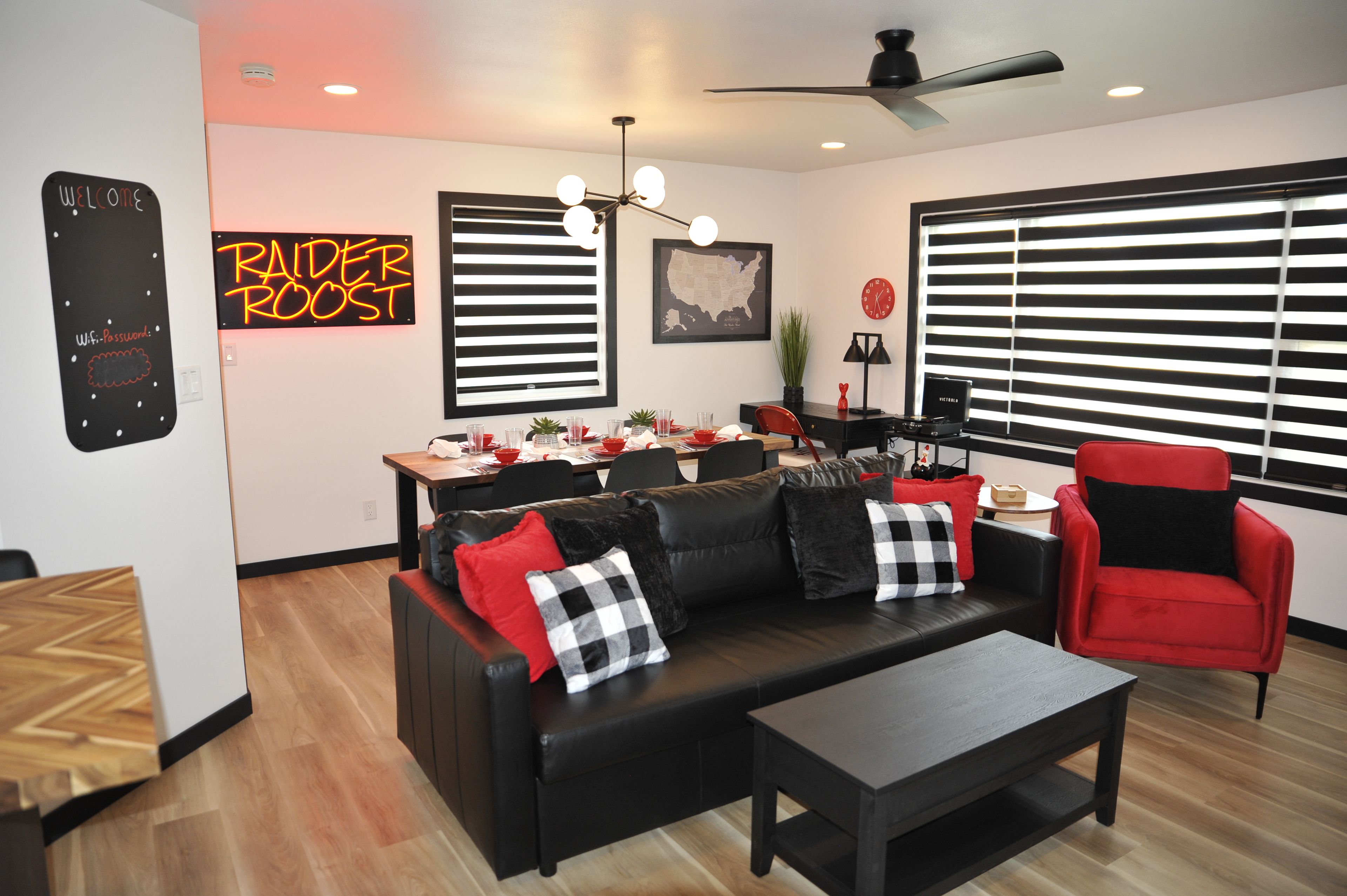 A modern living room with a black leather sofa, red and black cushions, a wooden coffee table, and a red armchair. There is a dining area with a wooden table and chairs, a map on the wall, and a ceiling fan.