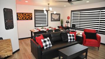 A modern living room with a black leather sofa, red and black cushions, a wooden coffee table, and a red armchair. There is a dining area with a wooden table and chairs, a map on the wall, and a ceiling fan.