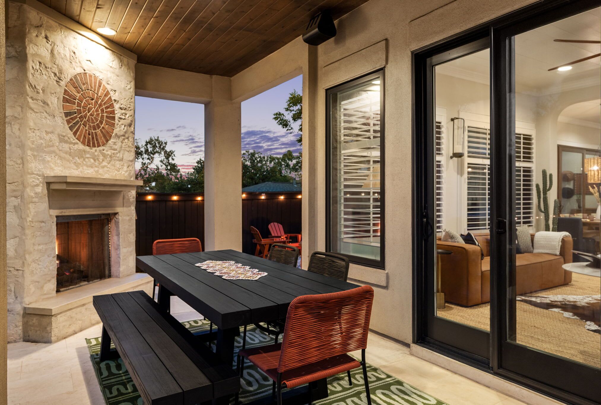Un patio al aire libre con chimenea de piedra, una mesa de comedor con sillas y un ventanal corredero que da a un espacio interior.
