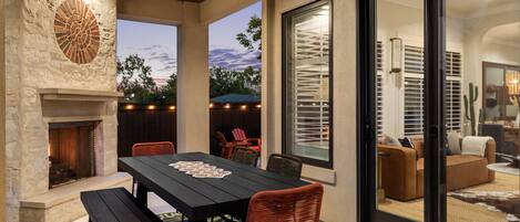 Un patio al aire libre con chimenea de piedra, una mesa de comedor con sillas y un ventanal corredero que da a un espacio interior.