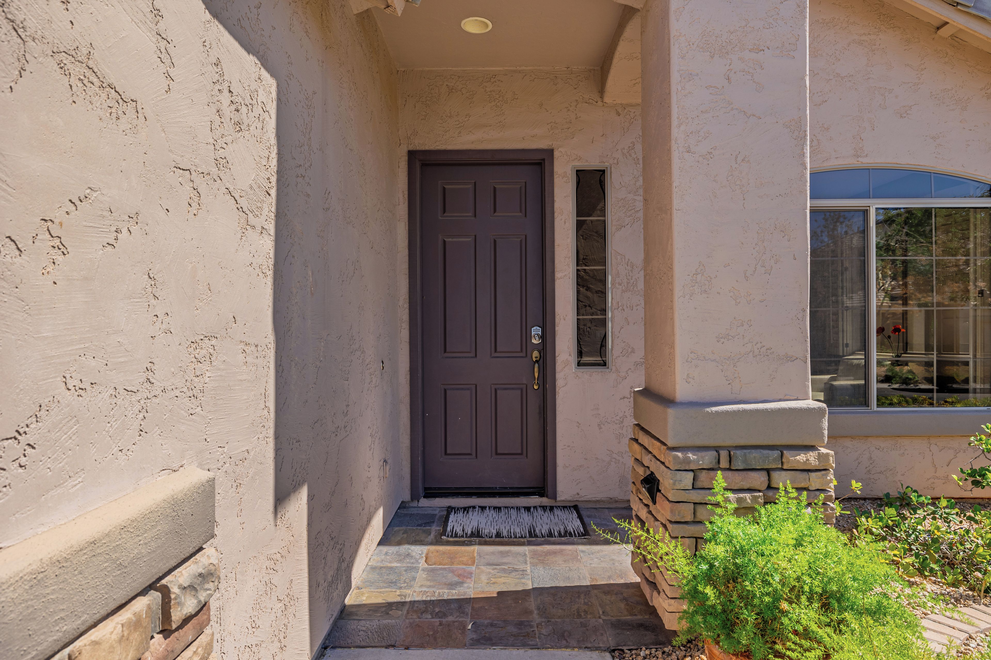 A house entrance with a dark door, a small porch with stone steps, and a window with a view of the outdoors.