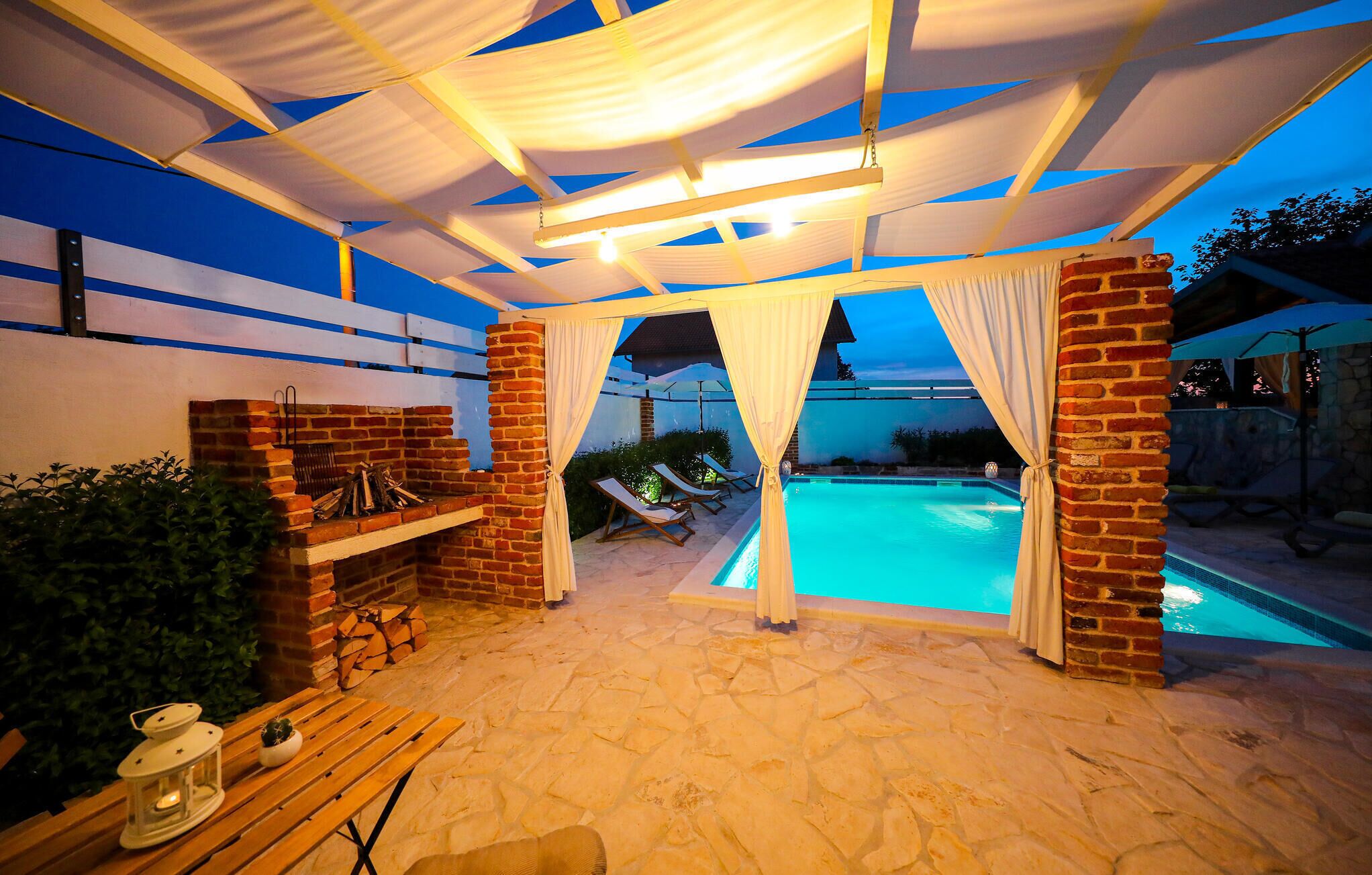 An outdoor pool area with a brick fireplace, lounge chairs, and a covered seating area with white curtains.