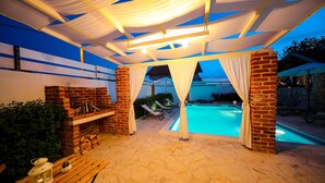 Un área de piscina al aire libre con chimenea de ladrillo, sillones de descanso y un espacio de estar cubierto con cortinas blancas.