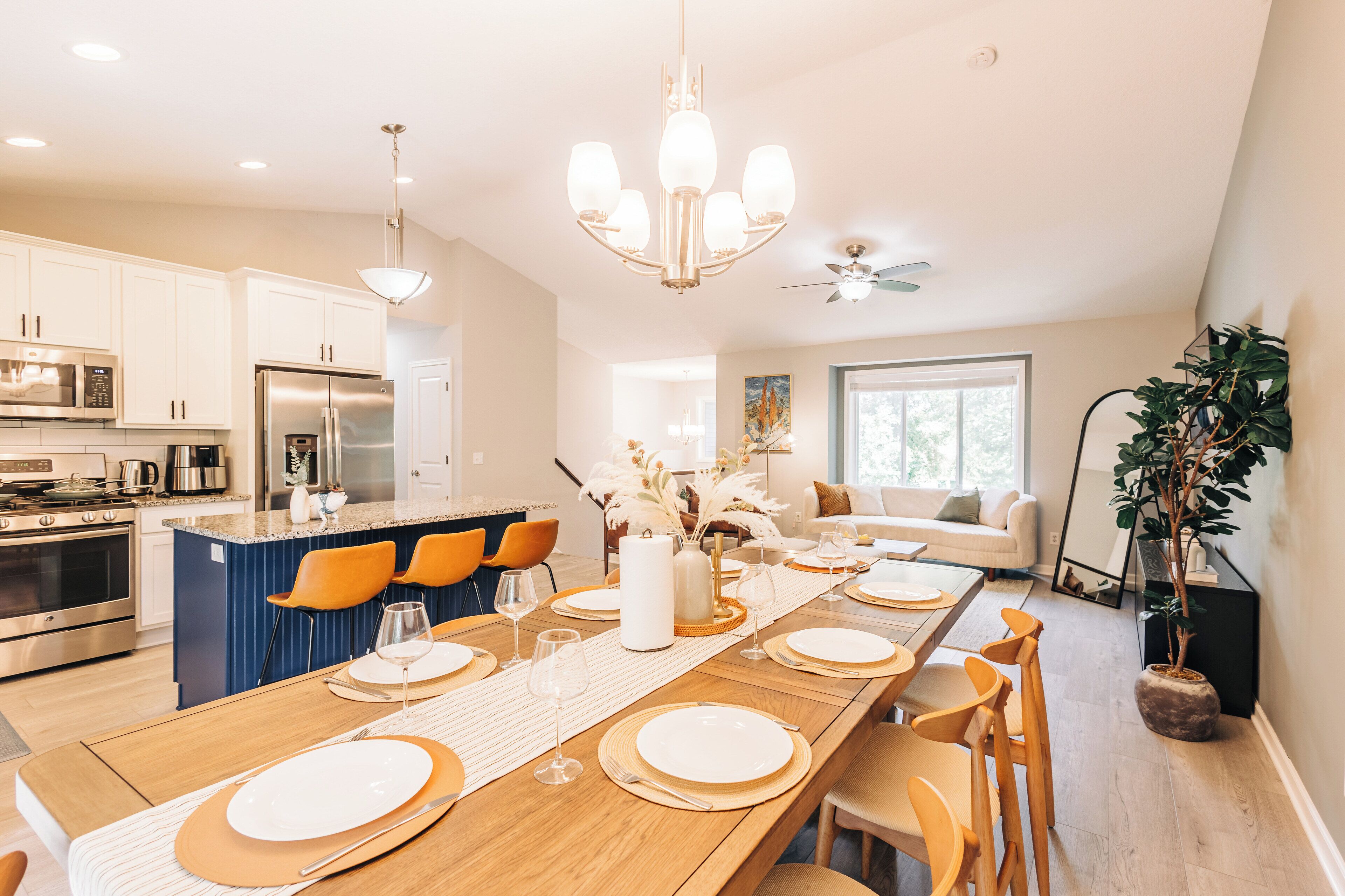 A modern kitchen with stainless steel appliances, a dining area with a long wooden table set for six, and a cozy living space with a sofa and a large window.