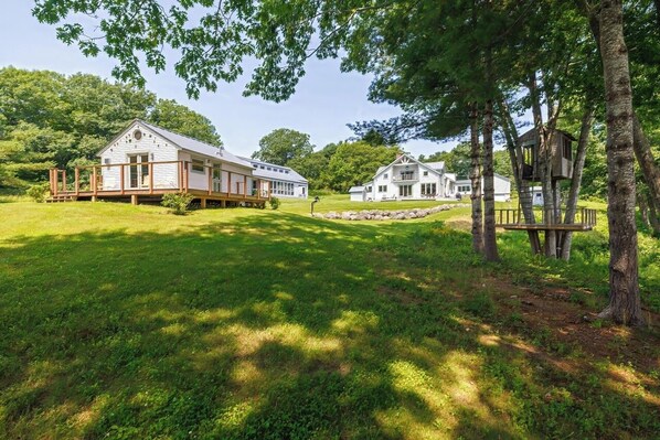 Une grande maison blanche avec une terrasse et une cabane dans les arbres, située dans un espace verdoyant.