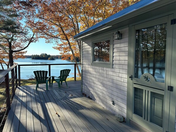 A wooden deck with two green Adirondack chairs facing a lake, a cabin with a glass door, and a metal railing.