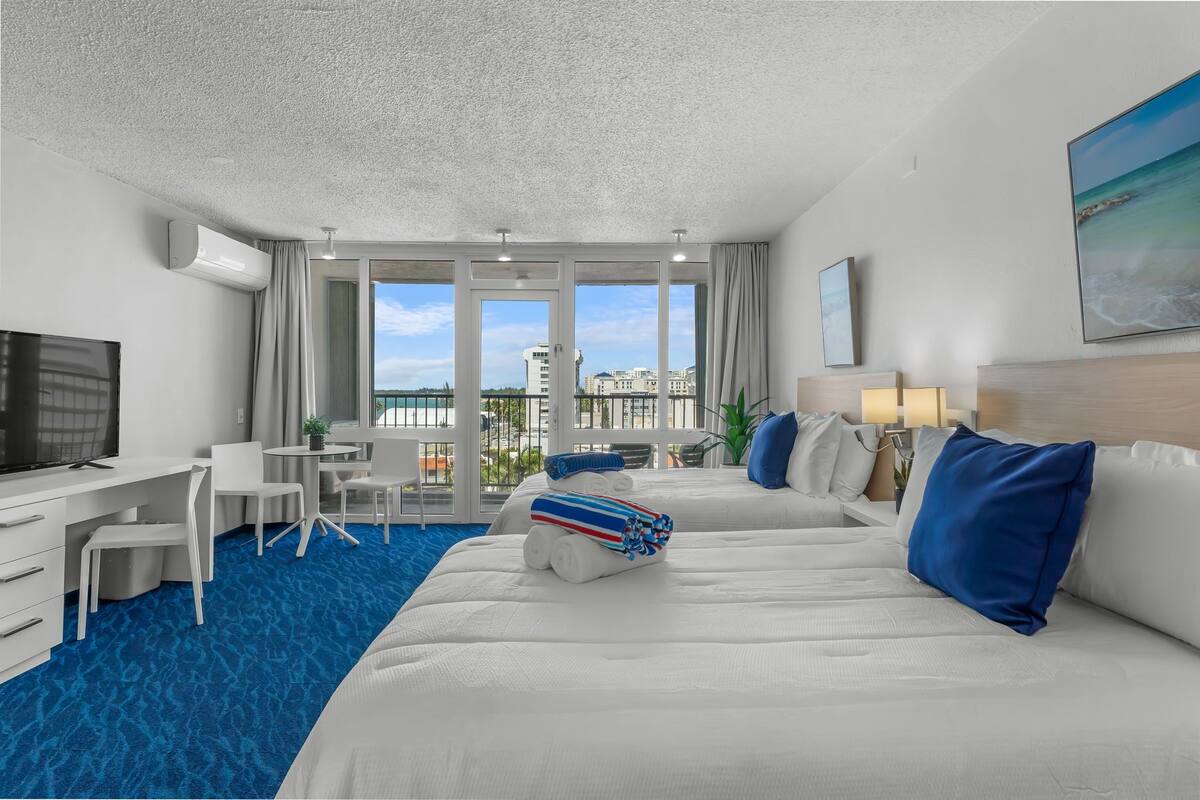 Ein Hotelzimmer mit einem großen Bett, einem Schreibtisch, einem Fernseher und einem Balkon mit Blick auf die Stadt.