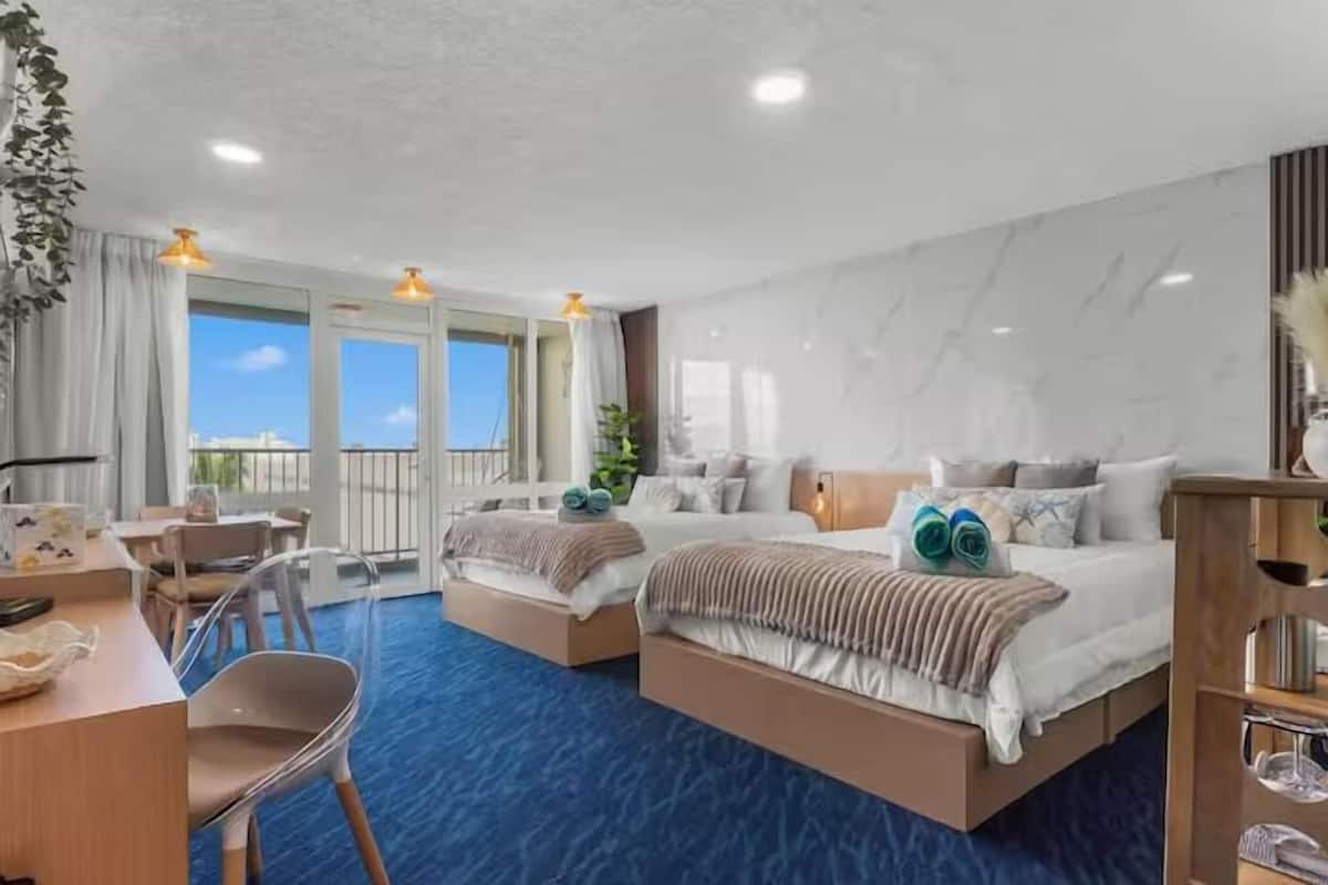 Ein Hotelzimmer mit zwei Betten, einem Schreibtisch und einem Stuhl. Es gibt einen Balkon mit Blick auf den Himmel.