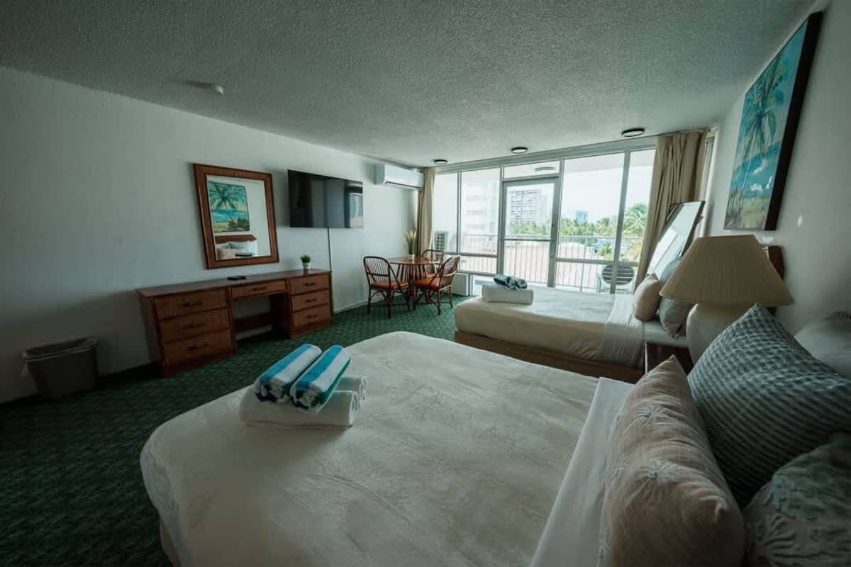 Ein Hotelzimmer mit einem großen Bett, einem kleineren Bett, einem Schreibtisch mit Stuhl, einem Fernseher und einem Balkon mit Blick.