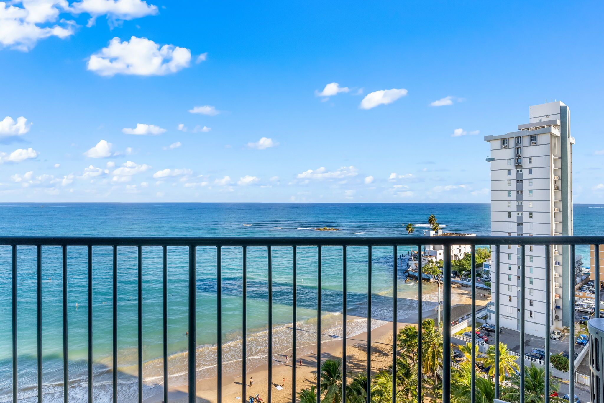 A coastal view from a balcony with a clear sky, a beach, and a tall building in the background.