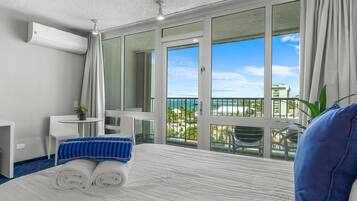A hotel room with a large bed, a small desk, a chair, and a balcony with a view of the ocean.