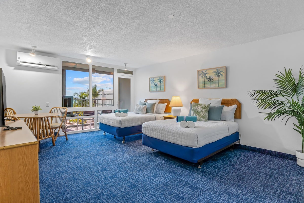 Ein Hotelzimmer mit zwei Betten, einem Schreibtisch und einem Balkon mit Blick auf Palmen.
