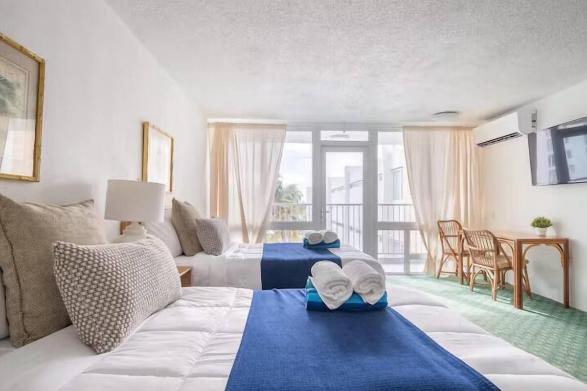 Ein Hotelzimmer mit zwei Betten, einem Schreibtisch, einem Sessel und einem Balkon mit Vorhängen.