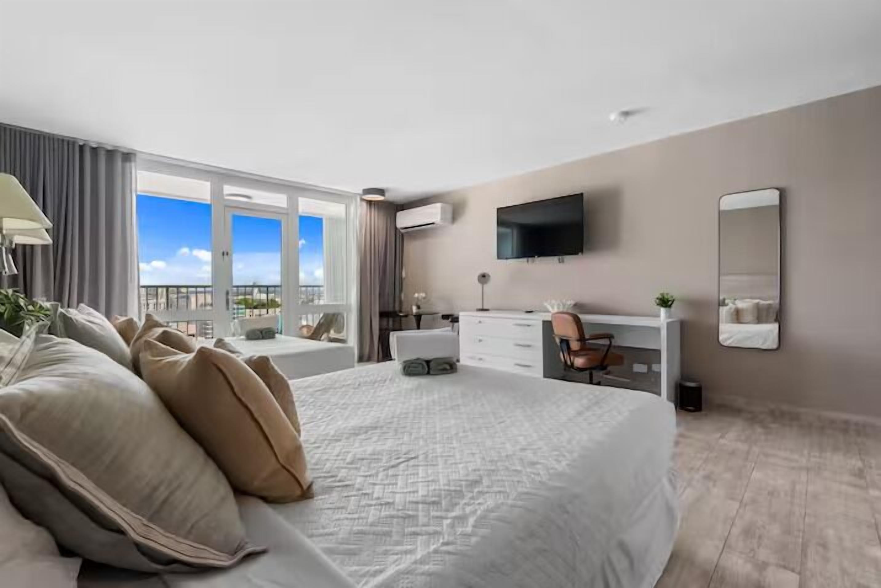 A modern hotel room with a large bed, a desk, a chair, a TV, and a balcony with a view.