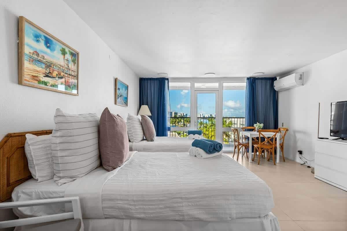 Ein Hotelzimmer mit zwei Betten, einem Esstisch mit Stühlen, einem Fernseher und einem Balkon mit Aussicht.