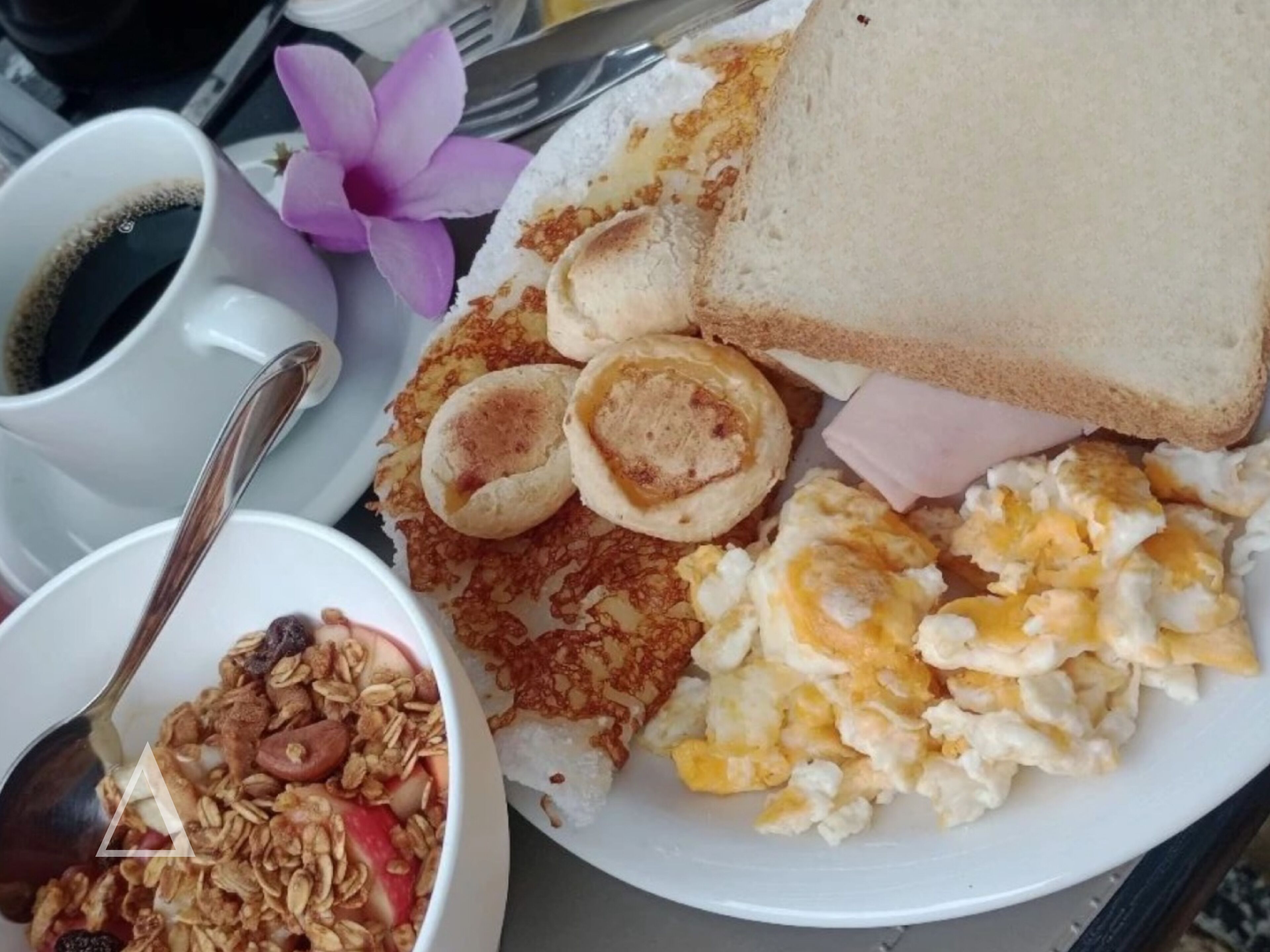 A breakfast plate with toast, eggs, ham, and pancakes, accompanied by a cup of coffee and a bowl of granola.