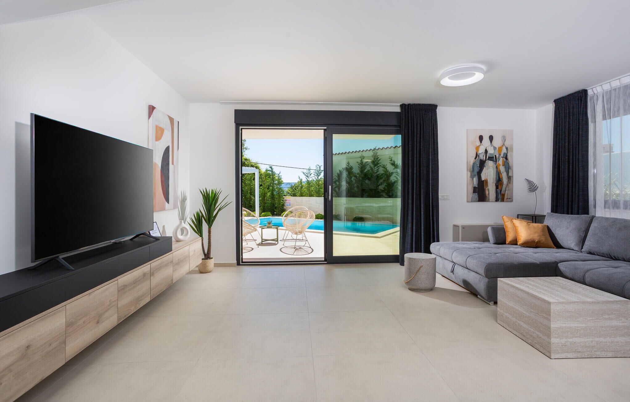 A modern living room with a flat-screen TV, a grey sofa, and a large sliding glass door leading to a pool area.