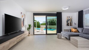 A modern living room with a flat-screen TV, a grey sofa, and a large sliding glass door leading to a pool area.