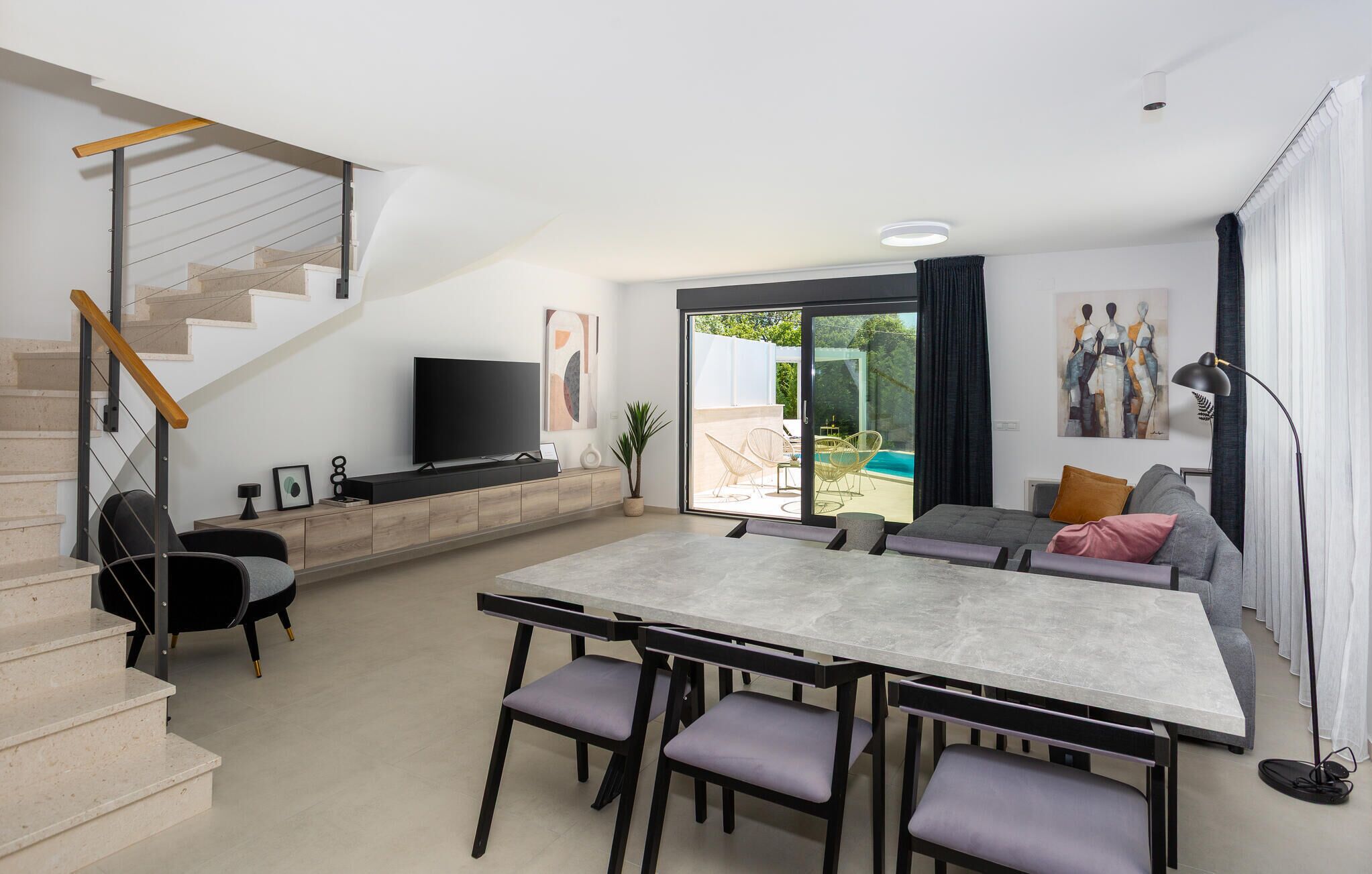 Una sala de estar moderna con una escalera, un televisor de pantalla plana, un comedor con una mesa grande y sillas, y un piso de vidrio corredera que da a un espacio exterior con piscina.