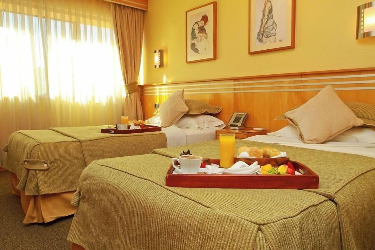 Habitación de hotel con dos camas, cada una con cubrecama verde, una mesita de noche con lámpara, una bandeja con artículos de desayuno y una obra de arte en la pared.