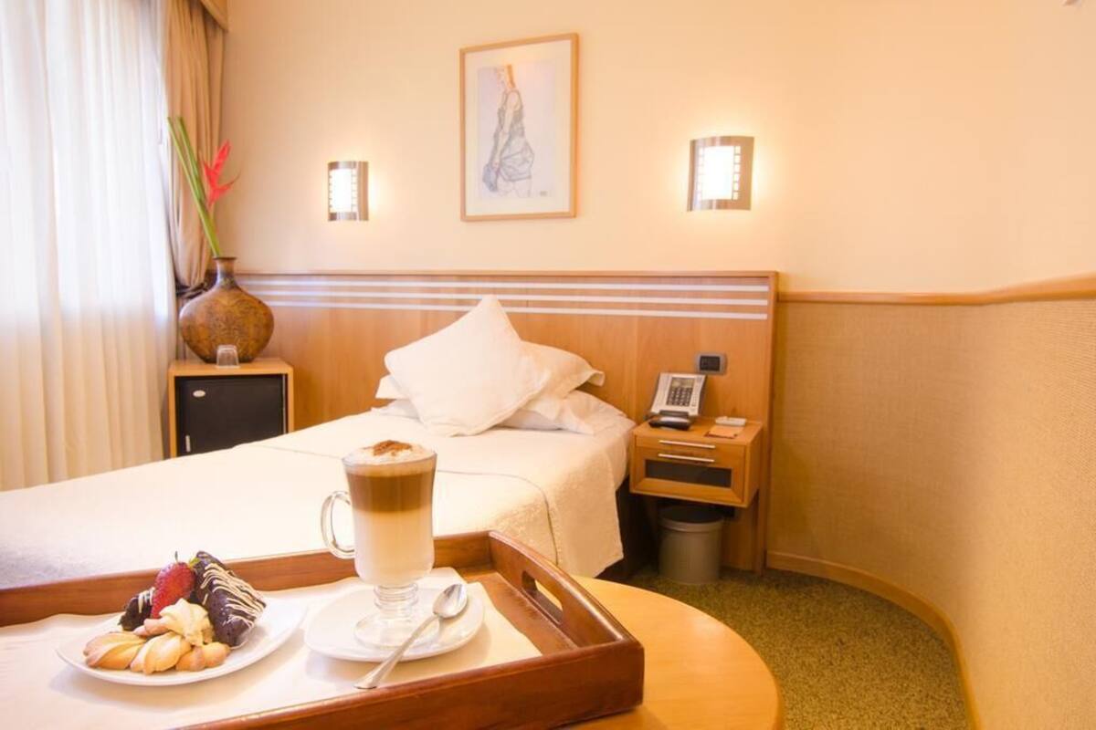 Habitación de hotel con cama, mesita de noche y bandeja de desayuno que incluye un plato de pasteles, una taza de café y un jarrón con flores.