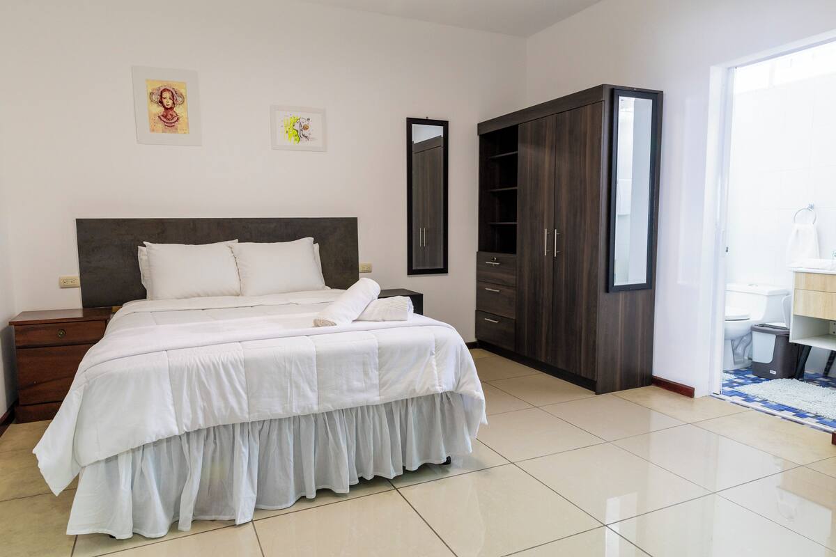 A bedroom with a bed, a wardrobe, a nightstand, and a bathroom visible through an open door.
