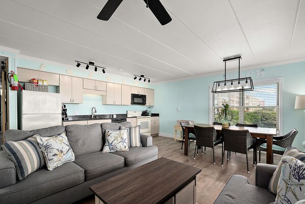 Una sala moderna con un sofá gris, una mesa de comedor y sillas. Se ve una cocina al fondo con armarios de color claro y un microondas.