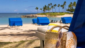 Une plage avec vue sur la mer, des chaises longues bleues et un sac en fibres naturelles posé sur le sable.