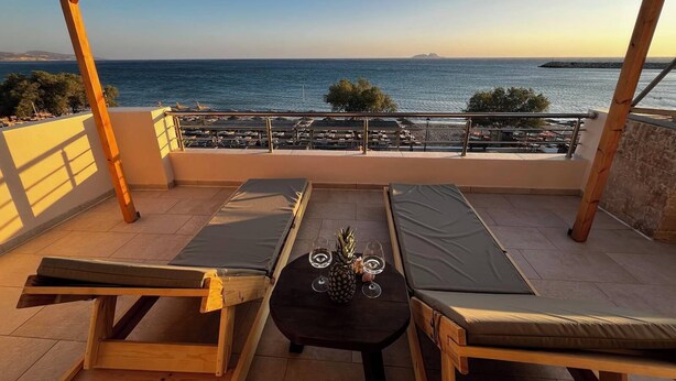 Un balcón con muebles de madera, una mesa con vasos y una piña, y vista al mar.