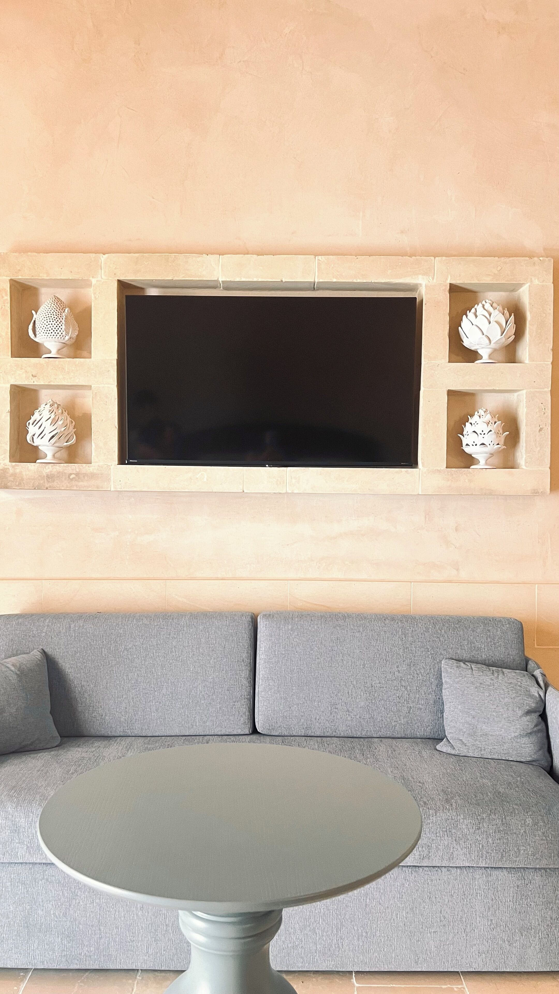 Un salón con un sofá gris, una mesa redonda y un televisor montado en la pared, junto con jarrones decorativos.