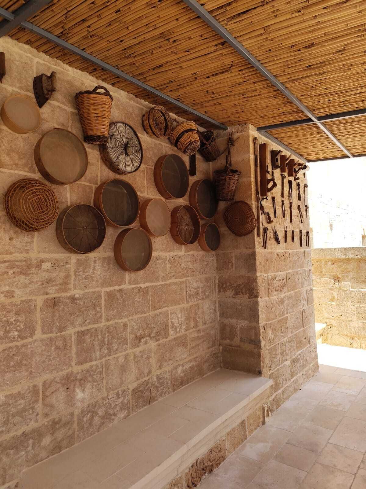 A stone-walled room with a thatched roof, featuring a variety of woven baskets and metal cookware hanging on the wall.