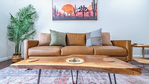 A living room with a brown leather sofa, a wooden coffee table with a coaster, a patterned rug, a large painting of a desert landscape, and a tall green plant. - 3 bed 2 bath house with 2 king beds, fire pit, gazebo & pool table! (Roswell)
