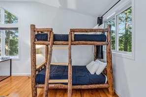 A bunk bed with a wooden frame and dark bedding, positioned in a room with large windows and a wooden floor.