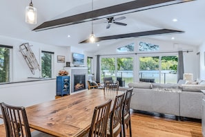 A spacious living room with a wooden dining table, comfortable seating, a fireplace, and large windows offering a view of the outdoors.