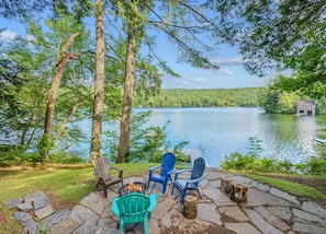 A lakeside deck with Adirondack chairs, a fire pit, and a view of a cabin across the water.