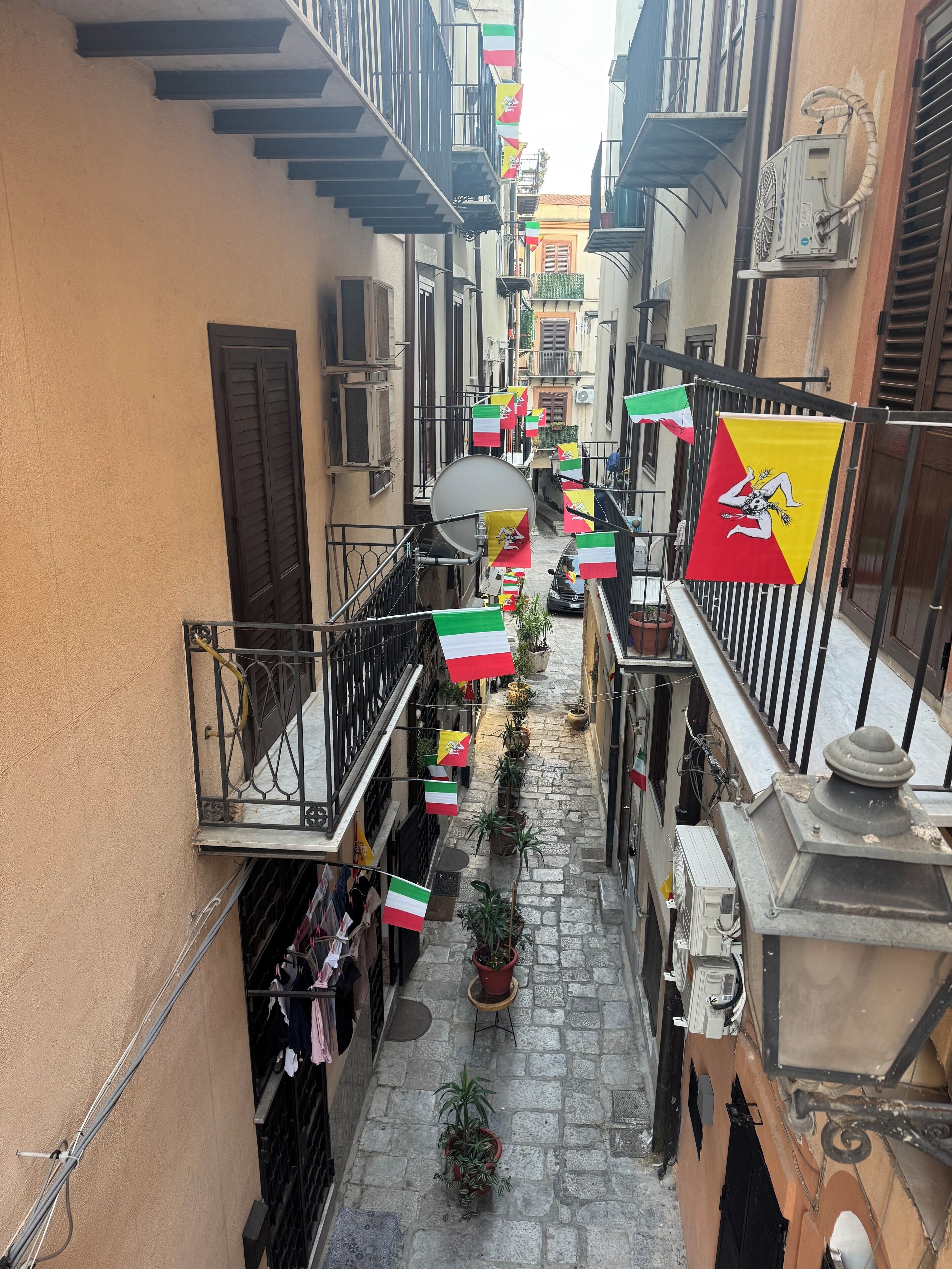 A narrow street with balconies, potted plants, and flags.