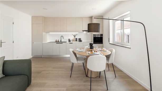 Une cuisine moderne avec des meubles blancs, une table à manger pour quatre personnes et une grande fenêtre qui laisse entrer la lumière naturelle.
