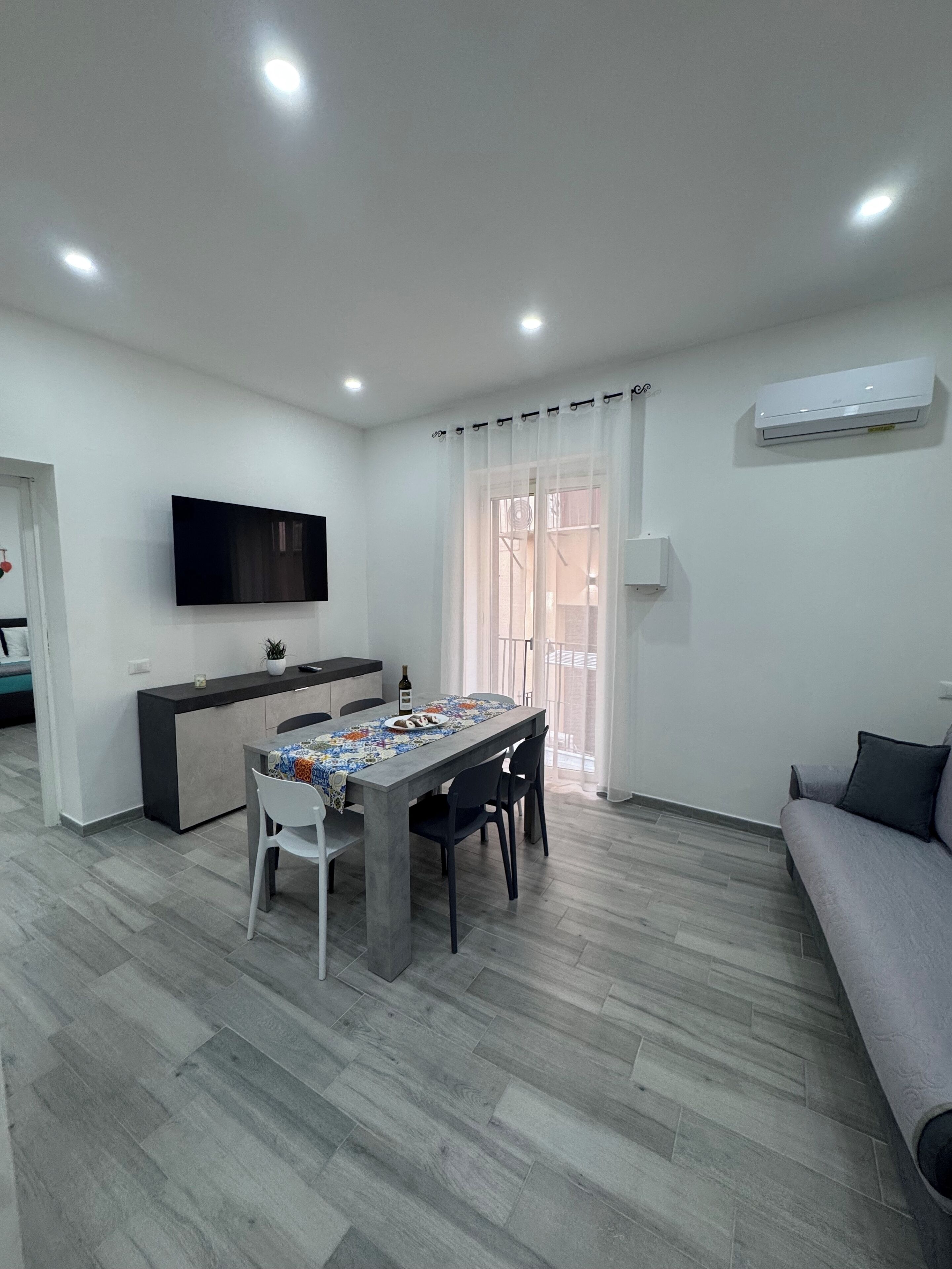 A modern living room with a dining area, a flat-screen TV mounted on the wall, and an air conditioning unit.