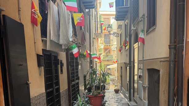 A narrow cobblestone street with potted plants and flags on the buildings. - Apartment in the historic center of palermo (Palermo)