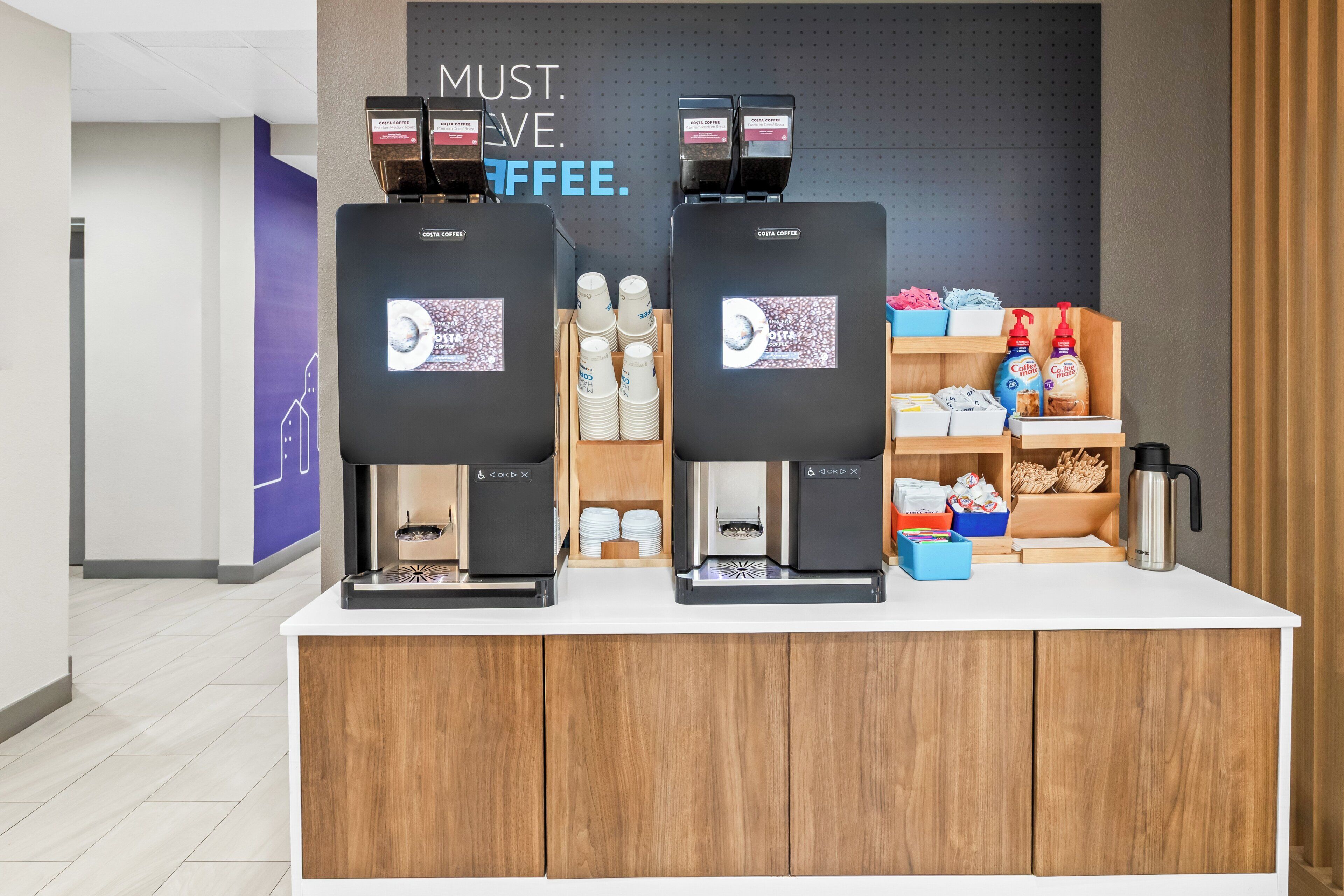 Una estación de café con dos máquinas de café, un cartel que dice 'MUST. LOVE. COFFEE.' y diversos artículos relacionados con el café.